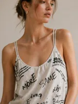 Blusa de tirantes finos dobles, color beige con estampado de letras y frases en color negro. Presenta un escote redondo y un diseño ligero.