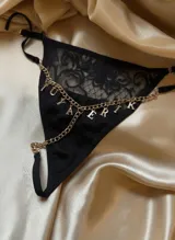 Tanga negra de encaje y lycra con tiras regulables y cadena dorada con dijes colgantes que forman letras.
