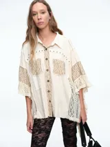 Camisa de corte oversize en color crudo, confeccionada con una mezcla de texturas que incluye paneles de encaje, detalles bordados y aplicaciones de tachas metálicas. Presenta mangas amplias tipo murciélago, cuello clásico y cierre frontal con botones.