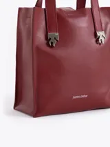 Cartera tipo tote pequeña de cuero color tostado, con doble asa superior y herrajes metálicos plateados en forma de llama en la base de las manijas. Presenta el logo de la marca estampado en la parte inferior frontal.