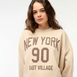 Buzo de cuello redondo en felpa color beige, con diseño estampado frontal con la inscripción 'NEW YORK 90 EAST VILLAGE' en color marrón. Presenta corte holgado y ruedo con terminación al corte.