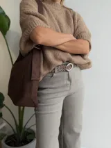 Bolso tipo bandolera de tamaño mediano, confeccionado en material con acabado efecto gamuza color chocolate. Presenta un diseño minimalista con cierre superior de cremallera y correa larga regulable con herrajes metálicos.