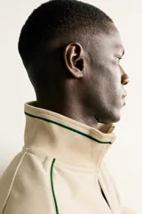 Chaqueta estilo deportivo de corte relajado, color beige con detalles de vivos en color verde oscuro. Presenta cuello alto, cierre frontal con cremallera de doble dirección, bolsillos laterales y puños elásticos.
