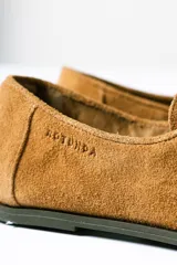 Mocasín de estilo relajado confeccionado en cuero gamuzado color tostado. Presenta detalles fruncidos en la puntera, cordones decorativos en el empeine y el logo de la marca grabado en el lateral.