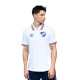 Remera tipo polo de Umbro del Club Nacional de Football, color blanco. Presenta cuello y bordes de mangas con detalles en azul y rojo, logo de la marca bordado en el pecho y escudo del club.