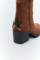 Bota corta estilo western de gamuza color marrón, con taco cuadrado negro y puntera estilizada. Presenta un detalle de estrella aplicada en el lateral y tirador en la parte superior.
