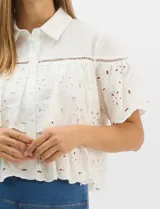 Camisa de algodón blanca con cuello clásico y cierre frontal de botones. Presenta un diseño con capas superpuestas y volados, combinando paneles de tela lisa con tejido bordado suizo de motivos calados y una cenefa decorativa. Posee mangas cortas y una silueta acampanada.
