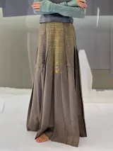 Falda larga plisada color marrón con estampado geométrico dorado en la parte superior y bolsillos laterales.