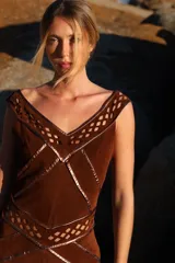 Vestido midi de gamuza color marrón cognac, con escote en V y detalles de calados geométricos artesanales en rombos. Presenta costuras decorativas con ribetes de cuero metalizado en tono cobre.