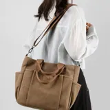 Bolso tipo tote bag de lona color negro, con múltiples bolsillos exteriores y asas cortas y correa larga de tela color marrón. Presenta un parche de cuero marrón con logo grabado en el frente.