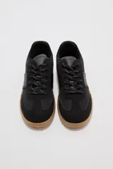 Zapatillas urbanas de color negro con suela de goma marrón, cordones negros con detalles blancos y paneles laterales.