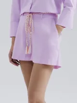 Short de lino color lila, de corte recto y tiro medio, con bolsillos laterales y cinturón trenzado ajustable con borlas en los extremos.