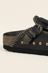 Zueco de cuero con diseño artesanal, presenta una tira con hebilla metálica decorativa, bordados sutiles y múltiples remaches metálicos en la parte frontal. El interior está forrado en cuero.