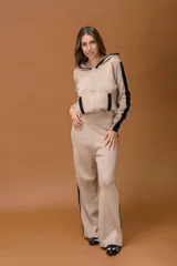 Conjunto de dos piezas en color beige con detalles en negro, compuesto por un buzo con capucha y cierre frontal, y un pantalón de corte recto y holgado. Ambas prendas presentan franjas laterales en contraste.