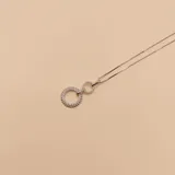 Collar de cadena tipo caja plateada con un dije compuesto por dos círculos entrelazados: uno más grande con borde de bolitas y pavé de circones, y otro más pequeño con pavé de circones.