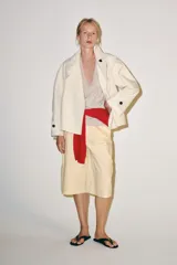 Trench corto color marfil de algodón, con cuello camisero, solapa cortaventos, mangas largas amplias con puños abotonados y cierre cruzado con un botón visible.