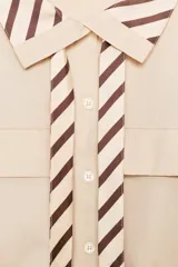 Camisa o cuerpo de manga larga color beige claro, con cuello solapa a contraste con estampado de rayas marrones y blancas. Presenta lazada a juego, bolsillos con solapa en el delantero y bajo con elástico.