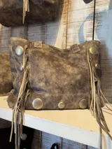 Bolso estilo boho de gamuza en tono grisáceo con efecto desgastado. Presenta flecos laterales largos, tachas metálicas circulares decorativas en los laterales y en la base, y costuras visibles. Diseño amplio con doble asa superior.