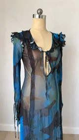 Vestido largo de microtul translúcido con estampado floral en tonos azules y marrones. Posee escote en V con lazo ajustable, mangas largas acampanadas con volados y una abertura lateral en la falda.