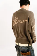 Sweater tejido reversible de corte boxy fit, con cuello redondo y puños acanalados. Presenta un diseño gráfico abstracto de estilo gótico que recorre el pecho y las mangas. Un lado es de color marrón oscuro con detalles en beige, y el otro lado es de color beige con detalles en marrón oscuro.
