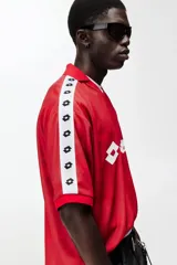 Camiseta de futbol de manga corta de la marca Lotto, en color rojo con un sutil estampado geométrico. Cuenta con cuello tipo polo abierto con escote en V y un calce holgado. Las mangas presentan una franja blanca lateral decorada con el logo de la marca repetido. El logotipo de Lotto destaca en blanco en el centro del pecho.