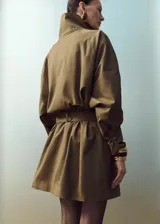 Chaqueta de gabardina color verde militar, de corte oversize con cuello alto abotonado de manera asimétrica, mangas largas con puños ajustables y cinturón ajustable del mismo tejido.
