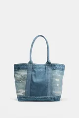 Bolso tipo shopper de hombro efecto denim, confeccionado con paneles de mezclilla de diferentes lavados y detalles deshilachados. Cuenta con doble asa de hombro y bolsillo interior con cremallera.