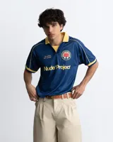 Remera de fútbol estilo vintage de la marca Nude Project, en color azul marino con cuello polo y detalles en amarillo. Presenta inscripciones y un escudo bordado en el pecho.