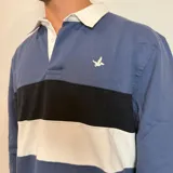 Buzo tipo polo de rugby con cuello blanco, manga larga y puños acanalados. Presenta un diseño de bloques de color en azul marino y verde, con un pequeño logo bordado en el pecho.