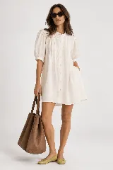 Vestido corto de lino natural, con cuello mao, mangas cortas abullonadas, cierre frontal con botones y pliegues decorativos en el pecho.