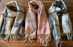 Bufanda larga de tejido suave con estampado a cuadros en tonos beige, gris, rosa y blanco, con flecos en los extremos.