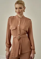 Camisa de gasa color capuchino, de manga larga con puños anchos abotonados, cuello mao y cierre frontal con botones dorados. Cuenta con bolsillos cargo en el pecho, detalles de charreteras en los hombros y un cinturón de tela ajustable con detalle de hebilla metálica dorada.