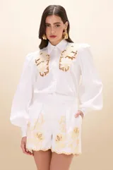 Camisa de tricolina color blanco, con cuello clásico, mangas largas abullonadas y puños ajustados. Presenta detalles de bordado calado con motivos florales en la parte superior delantera.