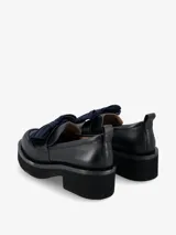 Mocasín de cuero negro con plataforma gruesa y taco cuadrado. Presenta un detalle decorativo de lazo plisado en color azul oscuro sobre el empeine y una pequeña tira en el talón.
