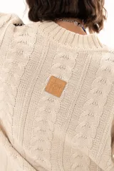 Buzo de tejido de punto color crudo con diseño de trenzas y costuras decorativas en color negro que recorren la prenda en forma de espiral. Presenta cuello redondo y mangas largas con terminación acampanada.