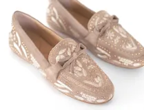 Mocasín de gamuza color avena con bordados florales en hilo blanco y pequeños apliques metálicos dorados. Presenta un lazo decorativo en la capellada y suela de goma flexible.