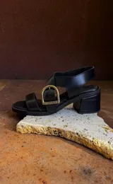 Sandalias de cuero negro con diseño de tiras anchas y hebilla metálica decorativa en tono bronce con el logo de la marca grabado. Presentan taco cuadrado de altura media y suela resistente.