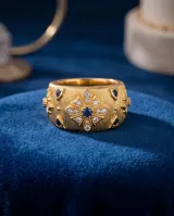 Anillo ancho de bronce con baño de oro, acabado texturizado y diseño con flor central incrustada de circonias blancas y una circonia azul en el centro.