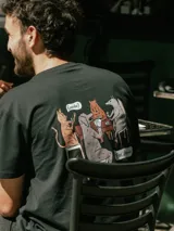 Remera negra de manga corta con estampado en la espalda que ilustra a varios animales jugando al truco con globos de texto que dicen 'Envido?' y 'Flor, atrevida!'.