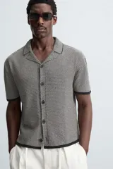 Camisa de punto de manga corta con cuello bowling y cierre frontal de botonadura. Presenta un patrón de rayas horizontales finas en blanco y negro, con ribetes negros en cuello, puños y bajo.