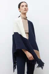 Ruana de tejido de punto con diseño color block en tonos crudo, gris y negro, con terminación de flecos en el ruedo y textura en relieve.