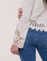 Cardigan corto beige de algodón con bordado en relieve, escote redondo y cierre frontal con botones carey. Mangas largas con detalles de encaje crochet y ruedo con terminación en ondas.