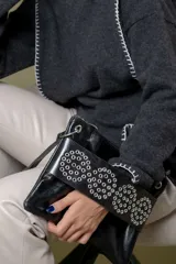 Sobre de mano confeccionado en cuero vacuno negro, con solapa decorada con múltiples remaches metálicos circulares. Cuenta con cierre principal y bolsillo trasero con cierre.