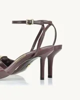 Sandalias de cuero color borgoña con taco fino y pulsera al tobillo. Presentan un aplique metálico dorado en forma de cuadrado en la parte delantera.