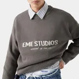 Suéter de punto grueso color gris oscuro, con corte cuadrado y ligeramente corto. Presenta cuello redondo, puños ajustados y el logo de la marca Eme Studios junto a la frase 'curated by ben ice' en jacquard blanco en el frente.
