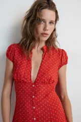 Vestido corto rojo con estampado de lunares blancos, cuello redondo con escote en pico pronunciado, manga muy corta y cierre frontal con botones.