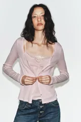 Chaqueta de punto color beige con escote en pico y manga larga, que incluye un top interior integrado con detalle de encaje a tono. Cierre frontal mediante botones.