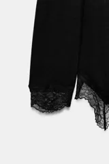 Top de manga larga en tejido suave color gris, con cuello redondo y bajo asimétrico decorado con encaje negro a tono. Presenta detalles de encaje negro en los puños.