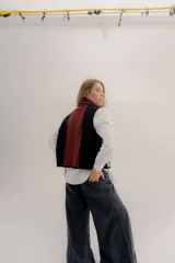 Chaleco abierto de tejido de punto, con diseño de bloques de color verticales en gris claro, negro y gris oscuro. Presenta un cuello ancho tipo chal y un detalle de tres pequeñas aplicaciones bordadas horizontales en tonos verde oliva, naranja quemado y celeste pálido en el frente.