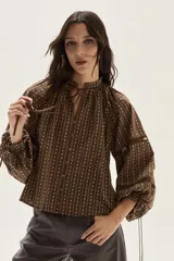 Blusa de color marrón con estampado de rayas verticales y pequeñas flores blancas. Presenta cuello redondo con lazo de ajuste, cierre frontal con botones y mangas largas abullonadas con detalles de encaje calado y puños ajustables con lazo.